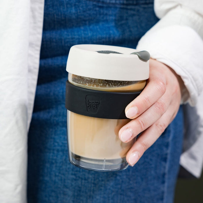 A hand holds a KeepCup Brew Longplay M 12oz in Filter, featuring a double-walled glass cup with creamy coffee inside, a black silicone band, and a light grey lid with dark grey plug, worn against a blue denim apron and white linen shirt.