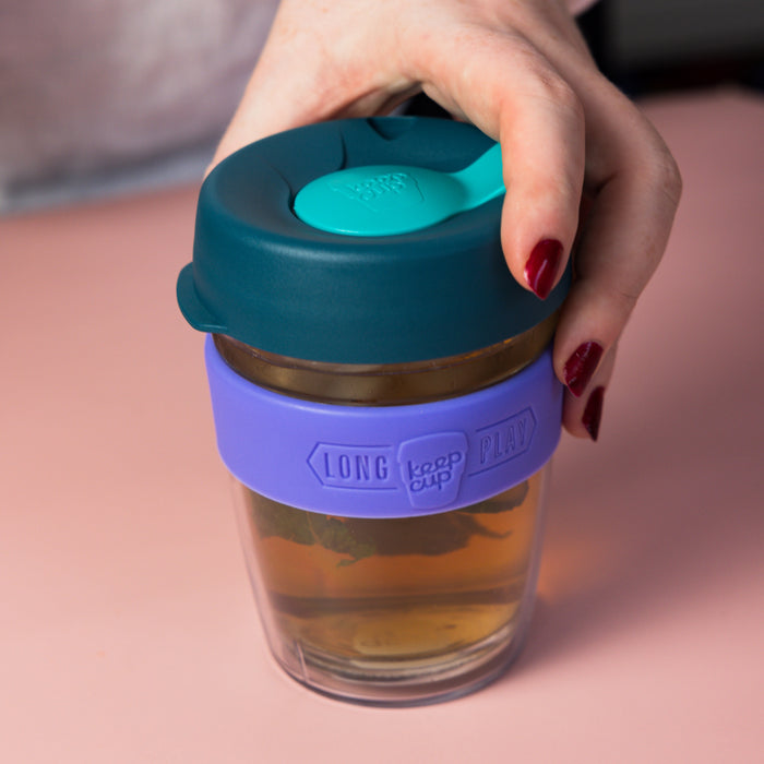 A hand with dark red nail polish reaches for a KeepCup Brew Longplay M 12oz in Haze, featuring a teal lid with turquoise sipper, purple silicone band, and double-walled glass cup containing tea, set against a soft pink surface.