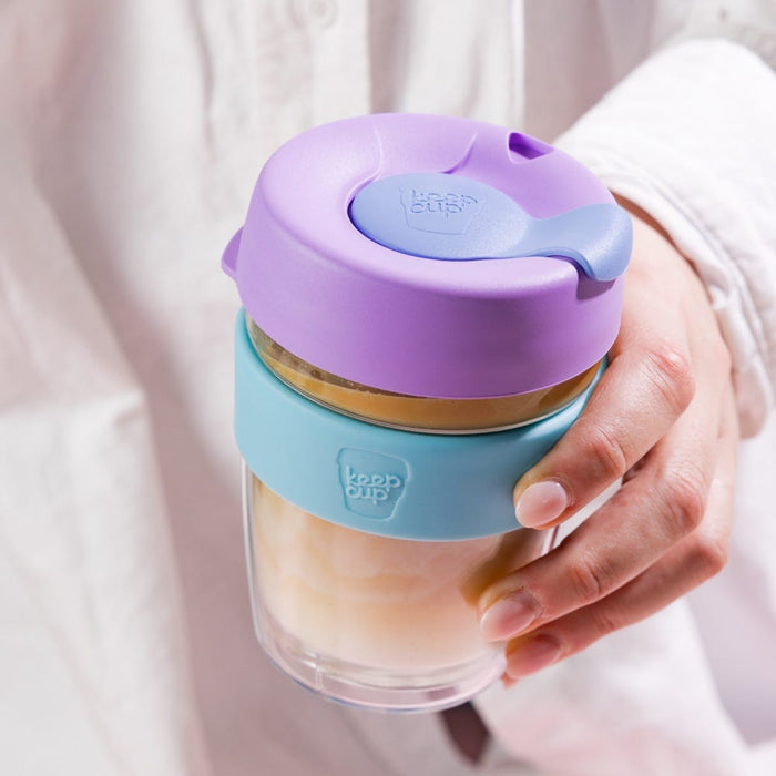 Hand holding a KeepCup Brew Longplay M 12oz in Moonlight, featuring a lavender lid with blue plug, aqua-coloured band, and double-walled glass cup filled with a creamy coffee drink.