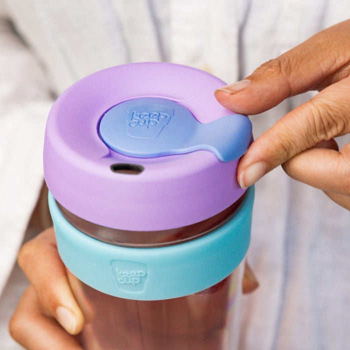 A hand holds the KeepCup Brew Longplay M 12oz in Moonlight, with fingers positioned to open the periwinkle blue sipper plug on the lavender plastic lid, showcasing the aqua-coloured band around the glass cup.