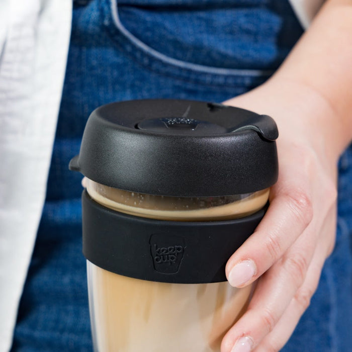 Hand holding a KeepCup Brew Longplay L 16oz in Black, filled with a creamy latte. The reusable glass cup features a black silicone band with embossed KeepCup logo and matching black lid, shown against a denim apron background.