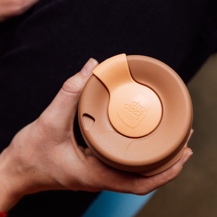 Hand holding a KeepCup Brew Longplay L 16oz in Canna, viewed from above showing the warm terracotta-coloured lid with the KeepCup logo on the rotating sipper closure.