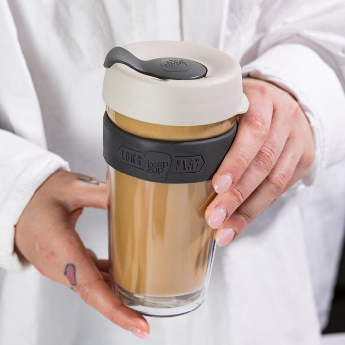 Hands holding a KeepCup Brew Longplay L 16oz in Filter colour, showing the double-walled glass body filled with a latte, charcoal silicone band with embossed branding, and cream-coloured lid with grey sipper plug.