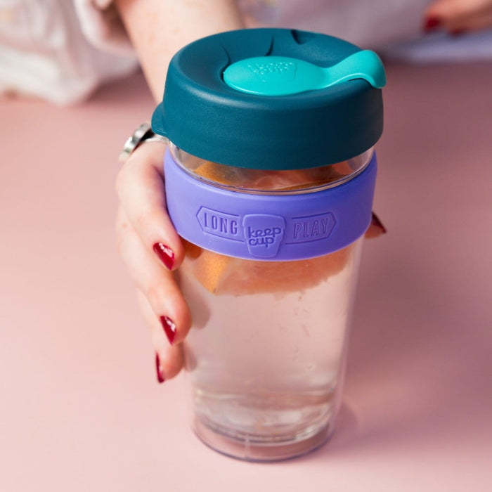 A hand with red nail polish holds the KeepCup Brew Longplay L 16oz in Haze, featuring a clear double-walled glass body with lavender silicone band, dark teal lid and turquoise sipper, filled with an iced beverage against a soft pink background.