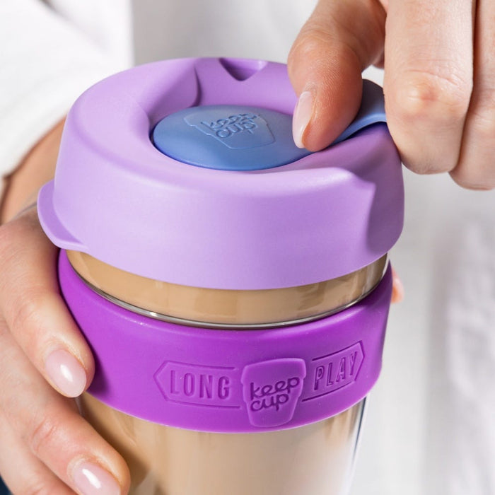 A hand holds the KeepCup Brew Longplay L 16oz in Helio, demonstrating the sipper plug on the lavender lid, while another hand grips the purple silicone band featuring the embossed "Long Keep Cup Play" branding, with creamy coffee visible through the glass body.