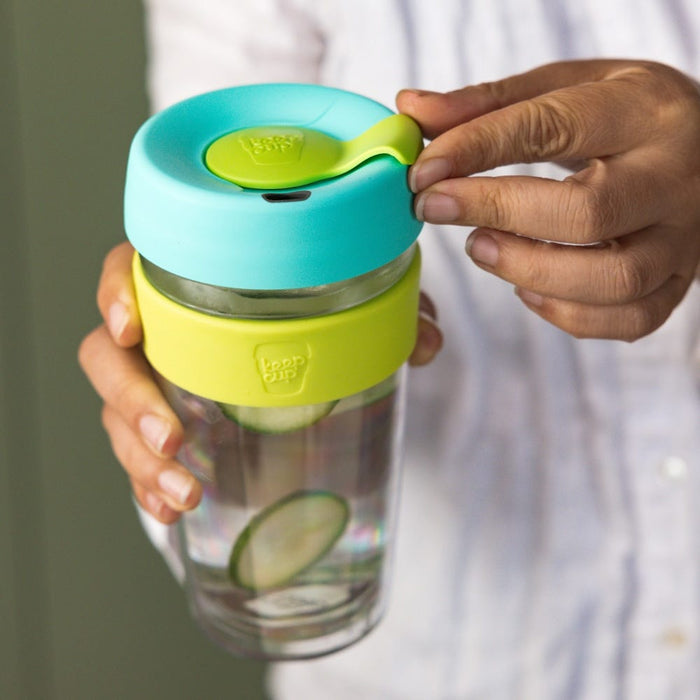 Hands holding a KeepCup Brew Longplay L 16oz in Matcha, featuring a turquoise lid with lime green plug and band, glass cup containing water with cucumber slices, with one hand opening the sipper plug.