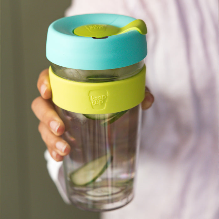 Hand holding a KeepCup Brew Longplay L 16oz in Matcha, featuring a clear double-walled glass body with lime green silicone band and turquoise lid, filled with water and cucumber slices.