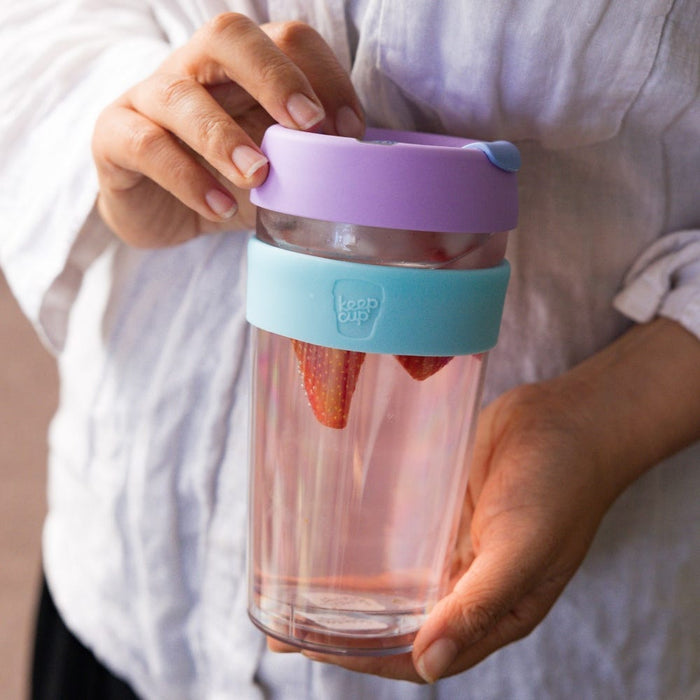 A person in a light grey linen shirt holds a KeepCup Brew Longplay L 16oz in Moonlight, featuring a clear double-walled glass body with pink-tinted water and fresh strawberries inside, a teal silicone band, and lavender lid with blue sipper plug.