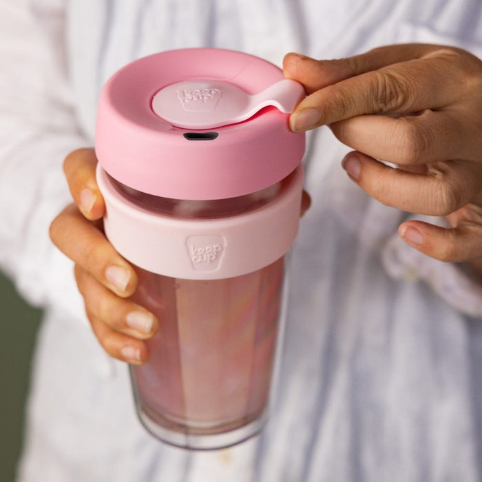 Hands holding a KeepCup Brew Longplay L 16oz in Pashmak Pink, with one hand gripping the clear glass cup with pink silicone band while the other lifts the pink lid's sipper closure.