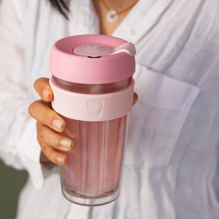 A hand holds the KeepCup Brew Longplay L 16oz in Pashmak Pink, showcasing the clear plastic cup with soft pink plastic lid and band, against a white linen shirt background.