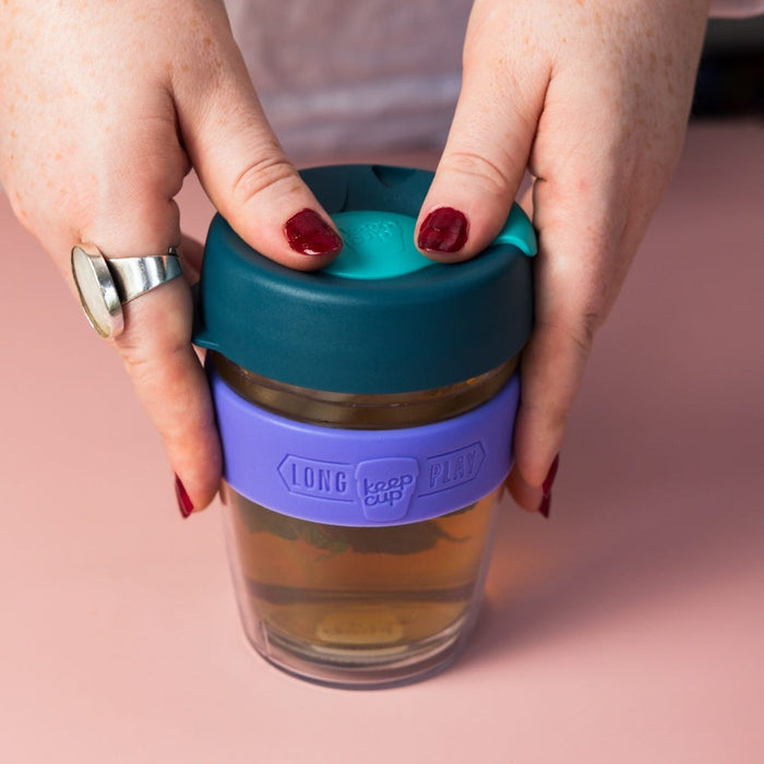 Hands with dark red nail polish holding a KeepCup Brew Longplay M 12oz in Haze, featuring a teal lid, purple silicone band with embossed "Long Play" and KeepCup logo, and double-walled glass cup containing tea, set against a pink background.