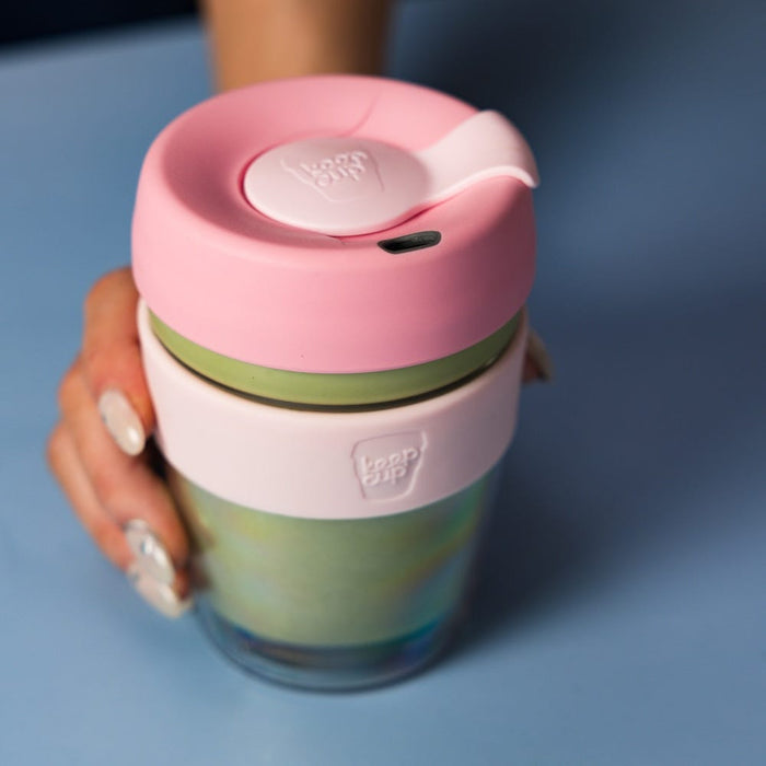 A hand with pale pink nail polish holds a KeepCup Brew Longplay M 12oz in Pashmak Pink, featuring a soft pink lid with flip-top closure, olive green band, and iridescent glass cup, shown against a muted blue background.