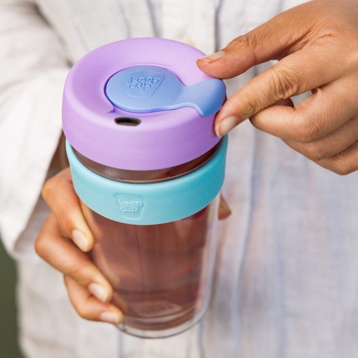 A hand holds the KeepCup Brew Longplay L 16oz in Moonlight, demonstrating the sipper lid closure with a finger pressing the blue plug tab. The reusable cup features a purple plastic lid, aqua band, and rose-tinted glass body.