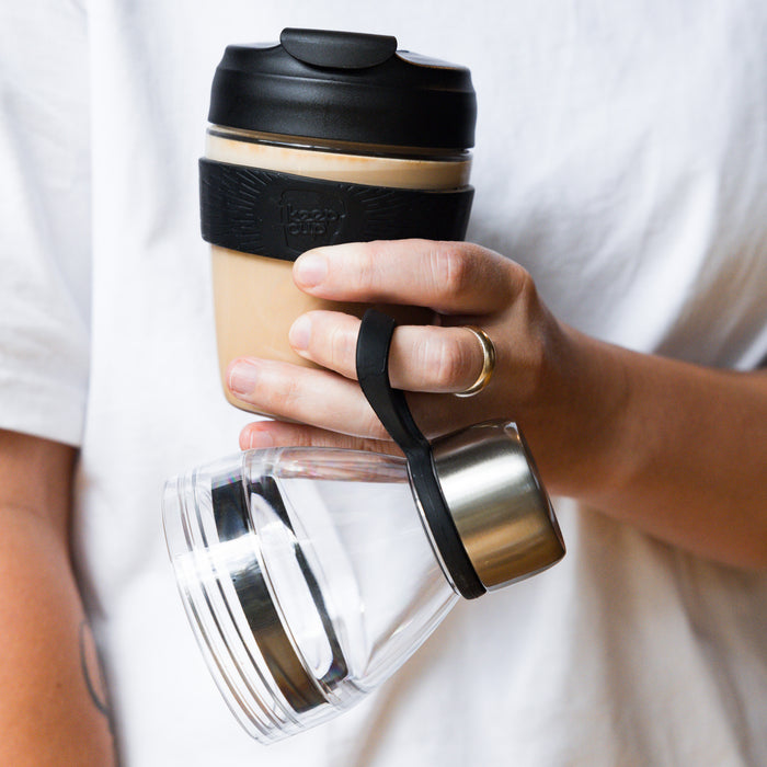A hand holds a KeepCup Glow Getter in Black, filled with a creamy coffee, while also carrying a clear double-walled glass carafe with a stainless steel lid by its black silicone strap, against a white shirt background.