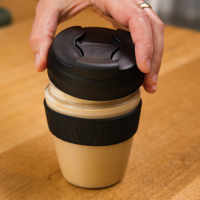 A hand reaches down to grasp the lid of a KeepCup Glow Getter in Black, featuring a cream-coloured glass cup with black plastic lid and band, placed on a wooden table.