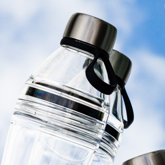 Close-up of the KeepCup Glow Getter in Black, shot from a low angle against a blue sky with soft clouds, highlighting the clear reusable bottle, stainless steel lid, and black carrying loop.