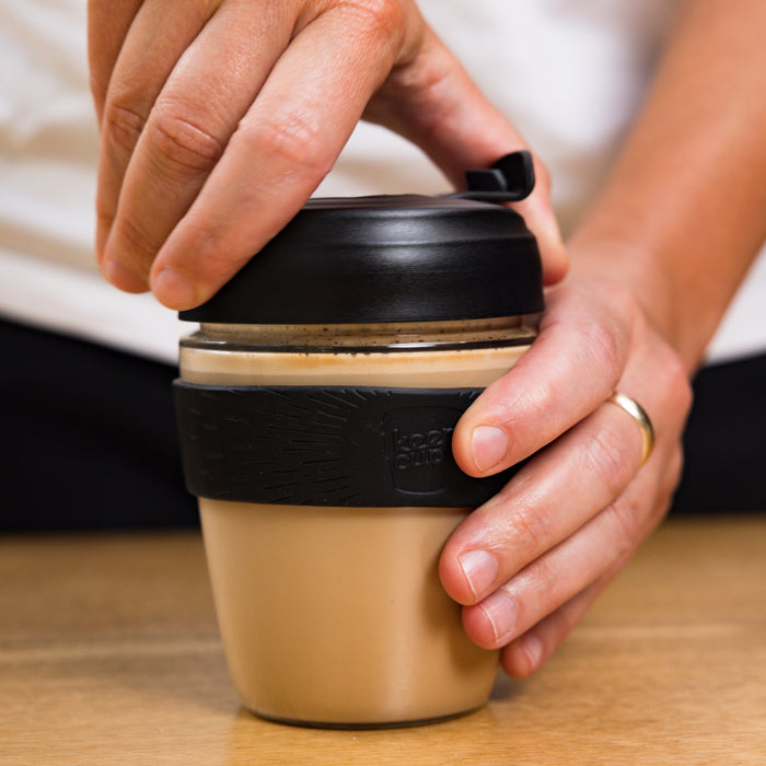 Hands holding a KeepCup Glow Getter in Black, with one hand on the lid and the other gripping the silicone band, showing the reusable cup filled with coffee resting on a wooden table.