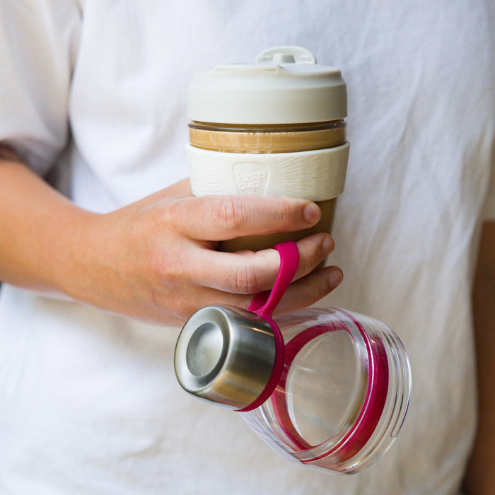 A hand holds a KeepCup Glow Getter in Burgundy, with the clear Tritan cup and magenta carry loop hanging from one finger, while the reusable coffee cup filled with a latte sits in the palm, featuring a cream-coloured lid and silicone band.