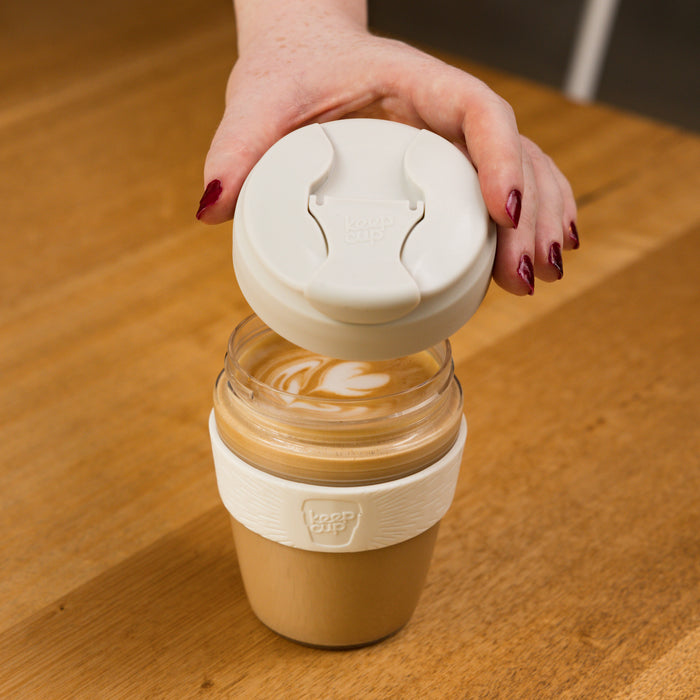 A hand with burgundy nail polish lifts the cream-coloured lid from a KeepCup Glow Getter filled with a latte featuring decorative milk art, set on a wooden café table.