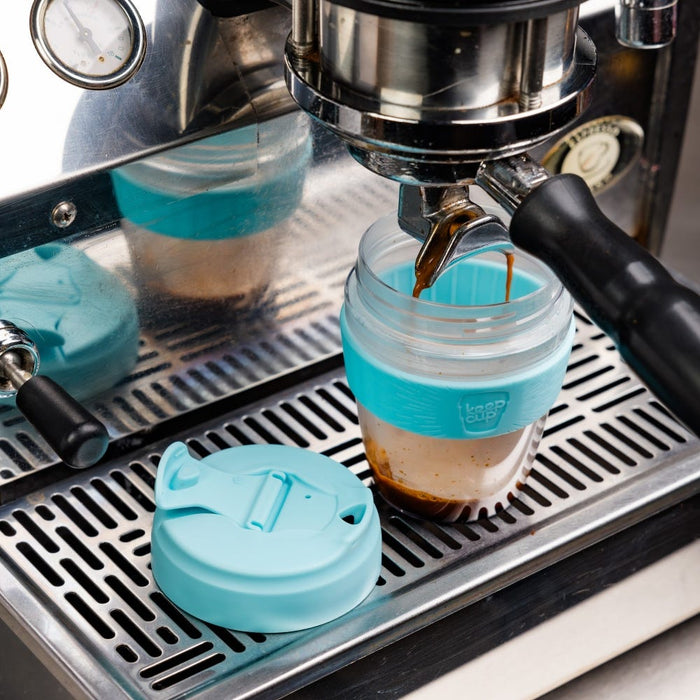 A KeepCup Glow Getter in Cloud colour sits on an espresso machine drip tray as fresh coffee pours into it from the portafilter, with the light blue plastic lid placed beside it and the cup's reflection visible in the machine's polished surface.