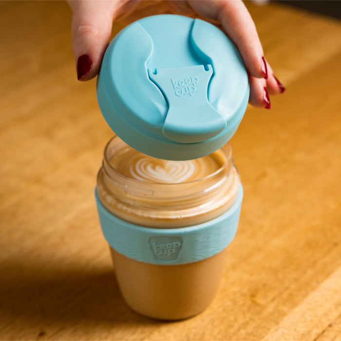 A hand with dark red nail polish lifts the light blue plastic lid from a KeepCup Glow Getter in Cloud, revealing a latte with heart-shaped latte art inside the glass cup with matching blue silicone band, set on a wooden table.