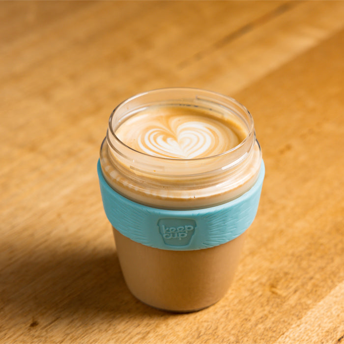 KeepCup Glow Getter in Cloud colour filled with a latte featuring heart-shaped latte art, photographed from above on a wooden table, showing the light beige cup body with pale blue silicone band.