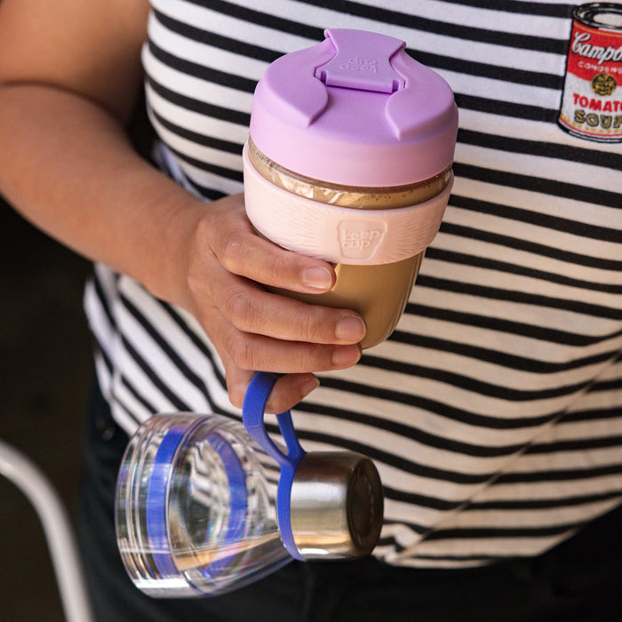 A hand holds the KeepCup Glow Getter in Lilac, featuring a lavender lid, pale pink band, and golden cork-coloured base, with a smaller clear glass KeepCup dangling from a blue loop below, against a black and white striped shirt background.