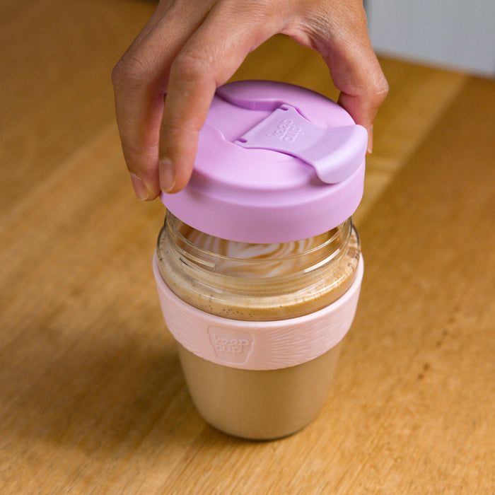 A hand lifts the lilac plastic lid from a KeepCup Glow Getter filled with a creamy latte, revealing the drink inside, set on a wooden table.