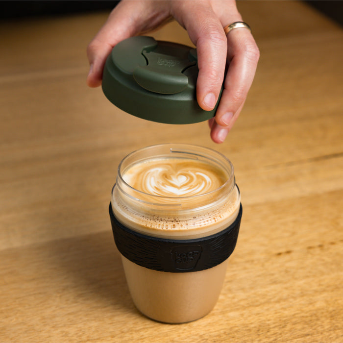 A hand lifts the pine green lid off a KeepCup Glow Getter reusable coffee cup, revealing a latte with heart-shaped latte art inside. The cup features a beige cork band with black silicone grip, sitting on a wooden table.