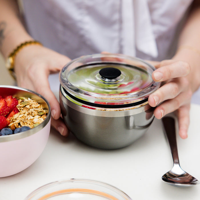 Hands holding a KeepCup Go Bowl Luxe in Nitro with flat lid, showing the stainless steel bowl filled with granola and fruit. A pink bowl with strawberries, blueberries and granola sits alongside on a white table with a spoon.
