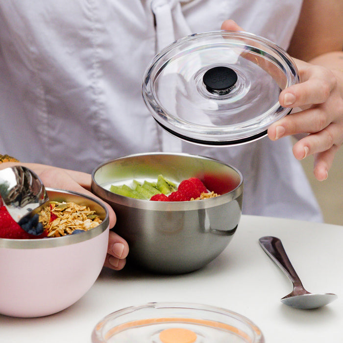 A person lifting the clear flat lid off a KeepCup Go Bowl Luxe in Nitro, revealing a stainless steel bowl filled with yoghurt, kiwi, raspberries and granola, while holding a pink Go Bowl in their other hand, on a white tabletop.