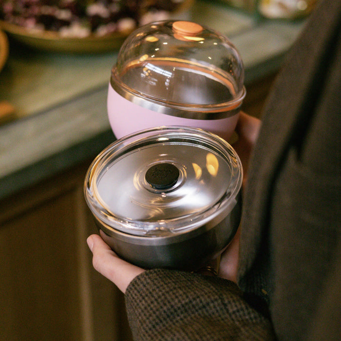A hand holds the KeepCup Go Bowl Luxe in Nitro with its flat lid, featuring a dark stainless steel base and clear domed lid, alongside a pink KeepCup in the background, both resting near a café counter.