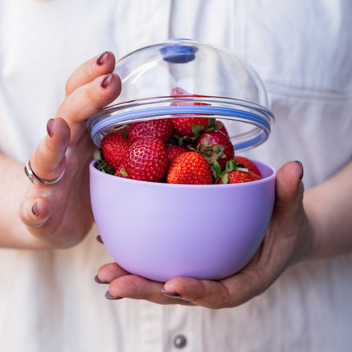 A person holds the Go Bowl Original M 550ml in Lilac, lifting its clear domed lid to reveal fresh strawberries inside the lilac coloured reusable bowl.