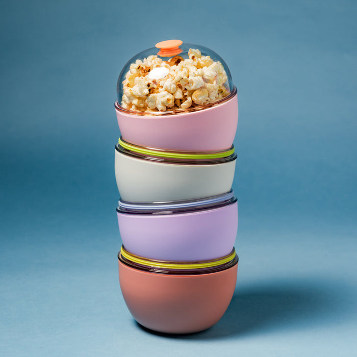A stack of four Go Bowl Original containers in different colours—clay, lilac, bone, and pink—with coloured  bands between each bowl, topped with a clear domed lid featuring an peach vent, the top pink bowl filled with popcorn, set against a blue background.