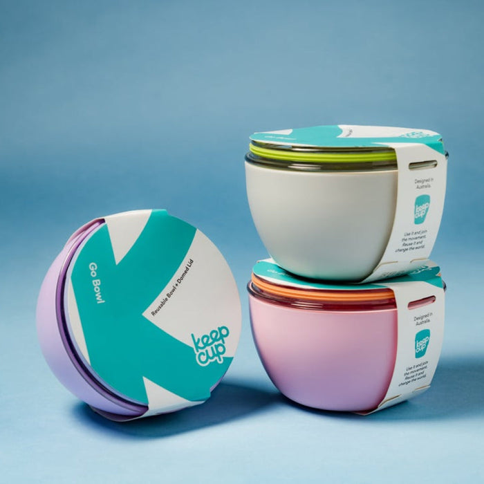 Three KeepCup Go Bowl Original reusable bowls with domed lids displayed against a blue background, featuring the Lilac colourway lying on its side in the foreground alongside stacked cream and pink variants, all with branded cardboard packaging sleeves showing the KeepCup logo and "Designed in Australia" text.