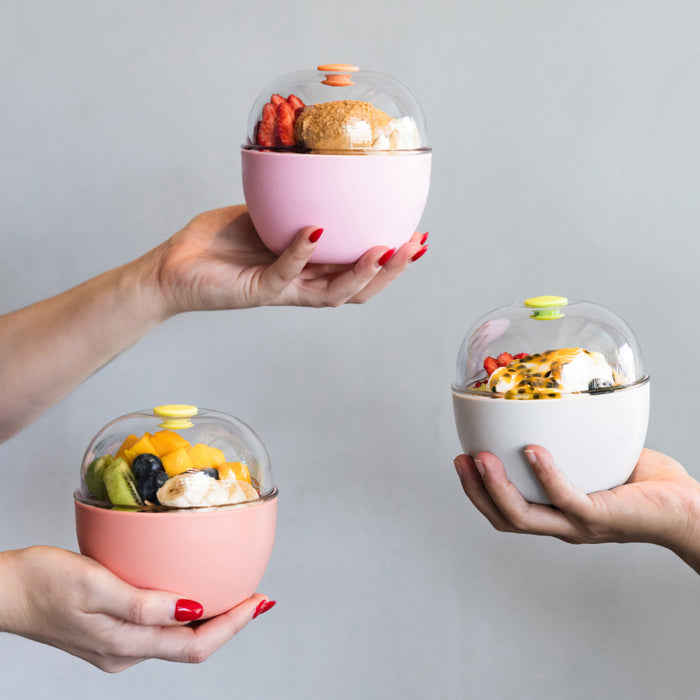 Three hands holding KeepCup Go Bowl containers in different colours—pink, clay, and the Go Bowl Original in Clay—each filled with yoghurt, fresh fruit, and toppings, showcasing the clear domed lid with colourful press-fit closures against a grey background.