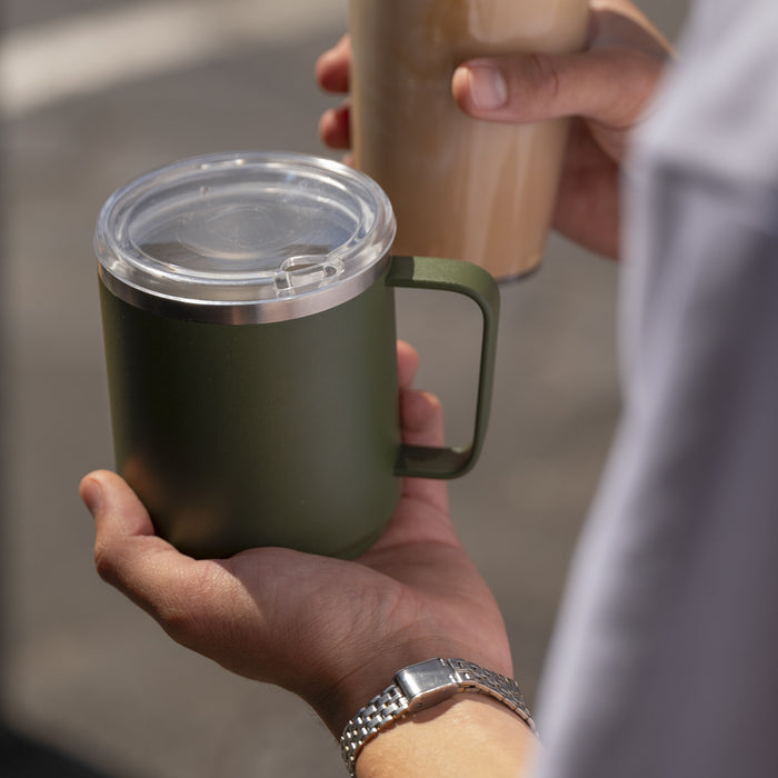 Person holding a green insulated mug with a clear lid and a beige cup in the background.