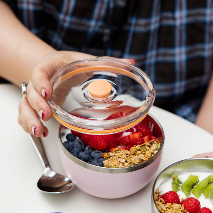 A hand with red nail polish lifts the clear plastic lid off a KeepCup Go High Go Low in Pashmak Pink, revealing a breakfast bowl filled with blueberries, raspberries, strawberries and granola, with a stainless steel spoon resting against the cup on a white table.