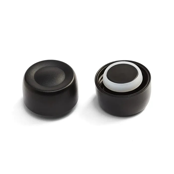 KeepCup Helix Bottle Cap in Black shown from two angles: the exterior view displaying the smooth matte black finish with concave top, and the interior view revealing the white silicone seal and ventilation slits.