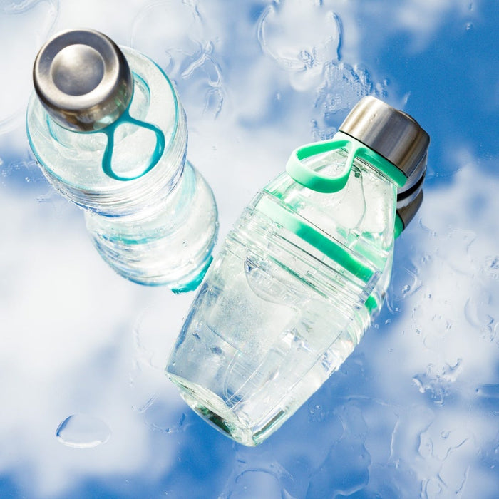 Two KeepCup Helix Light Bottles in Frost, featuring clear plastic bodies with mint green silicone carrying loops and stainless steel caps, lying on a wet surface with water droplets and a blue sky reflection.