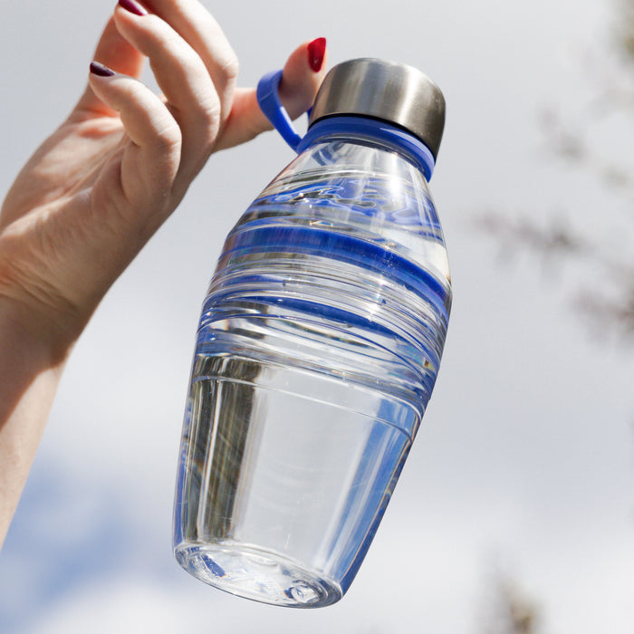 Hand with red nail polish holding a KeepCup Helix Light Bottle 530ml in Indigo by its carry loop, showcasing the clear tritan body with blue ridged grip band and stainless steel cap against a soft outdoor background.