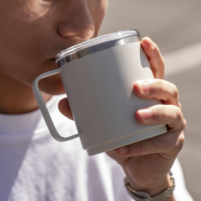A person in a white t-shirt drinks from a KeepCup Camp Mug M 12oz in Bone colour, holding the insulated stainless steel mug with both hands, showing the angular handle and clear sipper lid in natural daylight.