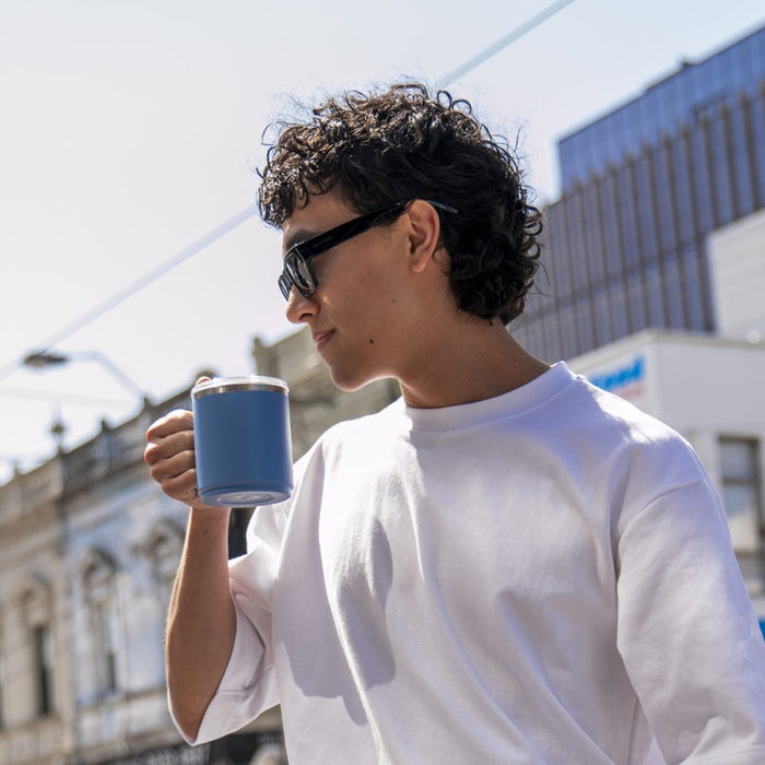 Young man with curly hair and sunglasses holds a KeepCup Camp Mug in Mist colour while walking through an urban streetscape on a sunny day.