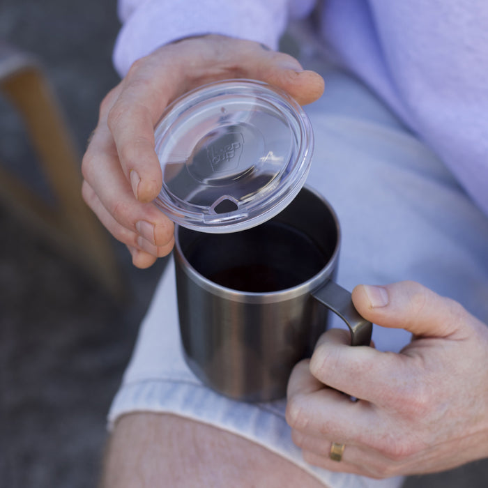 A person wearing a lavender jumper holds the KeepCup Camp Mug M 12oz in Nitro, lifting the clear plastic lid to reveal black coffee inside the stainless steel mug with handle.