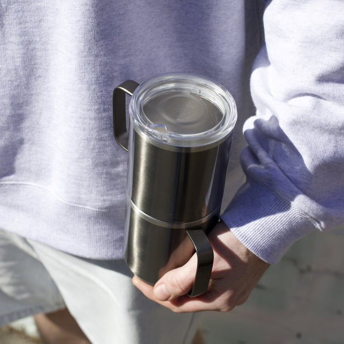 A person in a light purple sweatshirt holds a KeepCup Camp Mug M 12oz in Nitro, showcasing the brushed stainless steel body, sturdy handle, and clear press-fit lid in natural daylight.