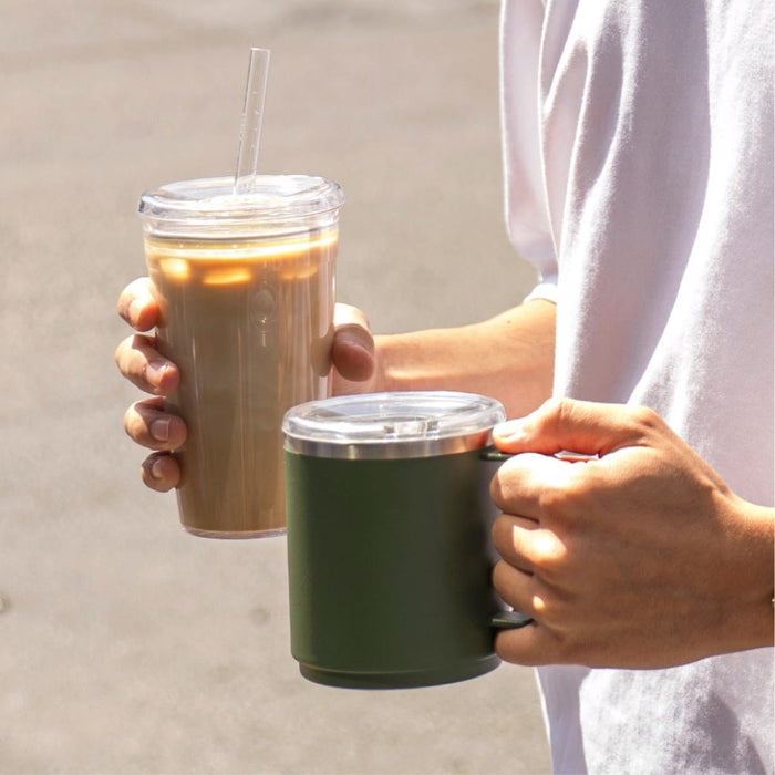 A person wearing a white t-shirt holds two reusable cups outdoors — a tall clear tumbler with iced coffee and straw in one hand, and the KeepCup Camp Mug in Pine with clear lid in the other, showcasing the mug's deep green colour and compact size.