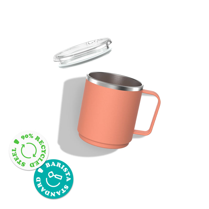 Camp Mug M 12oz in coral pink shown at an angle with clear plastic lid floating above, featuring stainless steel interior and matching handle. Badges indicate 90% recycled steel and Barista Standard certification.