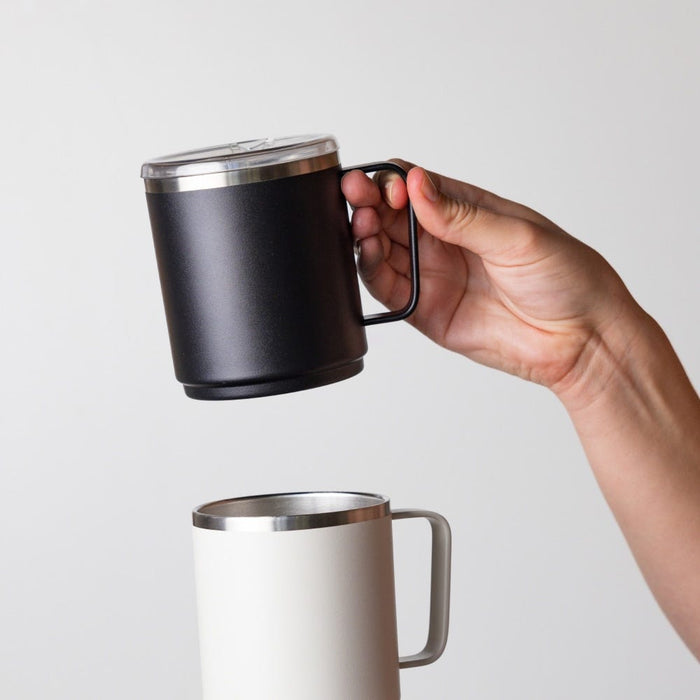 A hand holds the KeepCup Camp Mug M 12oz in Black above a white Camp Mug, showcasing the matte black finish, rectangular handle, stainless steel rim and clear sipper lid.