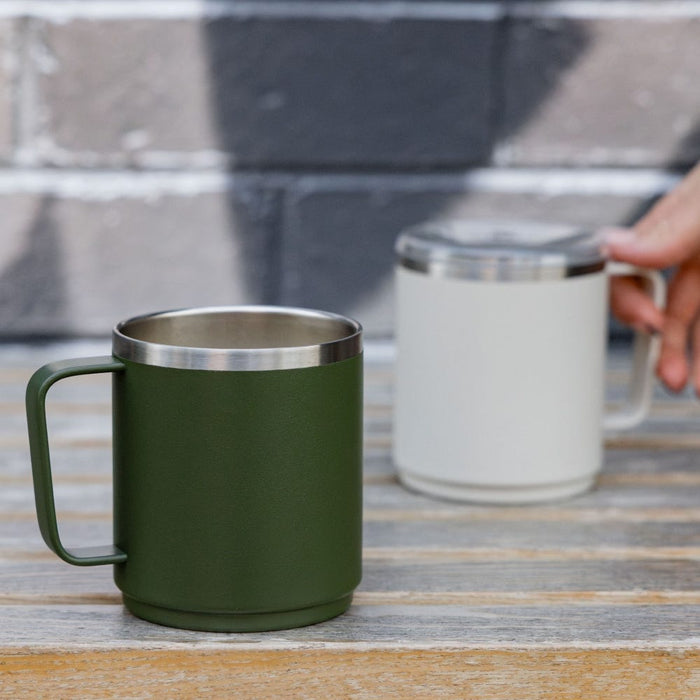 A dark green KeepCup Camp Mug M 12oz in Pine sits on a rustic wooden table in the foreground, with a hand reaching for a cream-coloured Camp Mug with lid in the background against a painted brick wall.