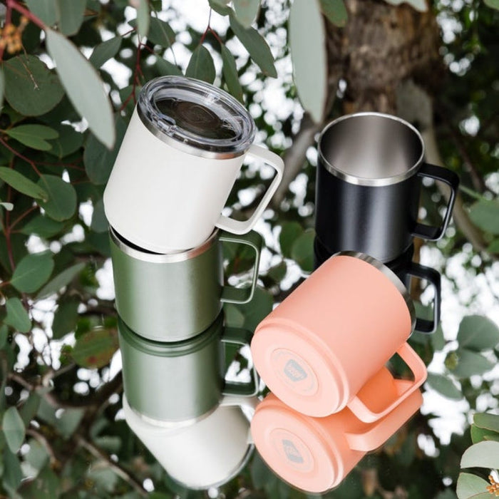 Four KeepCup Camp Mugs stacked and arranged outdoors among eucalyptus leaves, featuring the Bone colourway alongside Sage, Black and Coral options, showcasing the stainless steel interior and sturdy handle design.